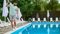 Man manually cleaning a swimming pool with a long skimmer pole beside lounge chairs and umbrellas at an outdoor pool area.