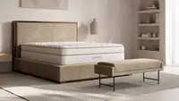 A modern bedroom featuring a white Saatva Classic mattress on a low-profile upholstered bed frame with warm wood accents, styled with neutral decor and soft natural lighting—an image highlighting the Saatva Memorial Day sale on luxury mattresses.