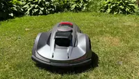 Silver Sunseeker Elite X7 robot lawn mower on grass in front of some bushes.