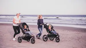 Two women jogging on a beach pushing kids in jogging strollers