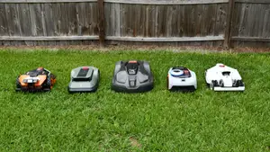 Five robot lawn mowers lined up together in the green grass with a fence behind