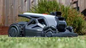 Grey and black AirSeekers Tron Robot Lawn Mower sitting on grass, in front of a wooden fence and bush.
