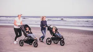 Two women jogging on a beach pushing kids in jogging strollers