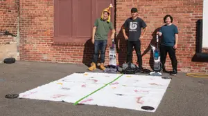 Three people stand in front of a brick wall and next to a white rug with stains on it.