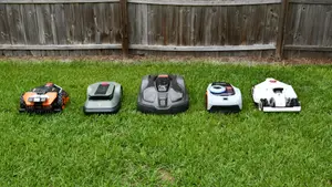 Five robot lawn mowers lined up together in the green grass with a fence behind
