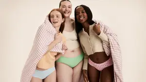 Three smiling women of diverse body types wearing period underwear and wrapped in a pink-striped blanket, posing together confidently.