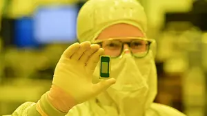 Person in a clean room suit holds a small computer chip up in gloved hand