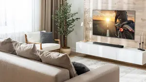 A bright, modern living room featuring an LG Dolby Atmos soundbar mounted beneath a large flat-screen TV on a light wood accent wall. The minimalist space includes a beige sectional sofa, white media console, indoor plant, and soft neutral decor, showcasing a clean and immersive home entertainment setup.