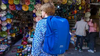A woman wearing a blue Osprey travel backpack stands outside a pottery vendor in a Morocco market.