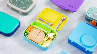 Bento boxes, cheese stick, carrots, apple, a sandwich, and M&Ms in a lunchbox, laid out on a marble counter.