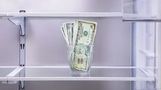 A glass with cash in it sitting on an empty refrigerator shelf.