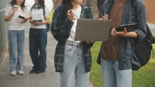 A group of students on the sidewalk holding notebooks, a laptop and an iPad