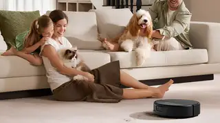 A woman and young girl sit on the floor with a Ragdoll cat while a man pets a fluffy dog on a cream sofa, as an iRobot Roomba robot vacuum cleans crumbs from the carpet beside them.