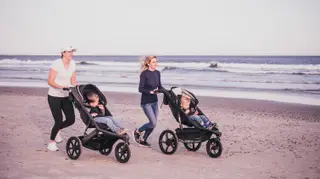 Two women jogging on a beach pushing kids in jogging strollers