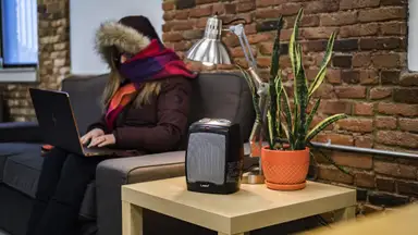 A woman sits on a couch in a cold room wrapped in a jacket.