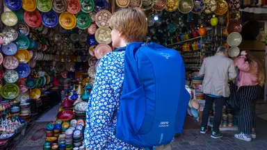 A woman wearing a blue Osprey travel backpack stands outside a pottery vendor in a Morocco market.