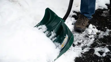 A close-up of a snow shovel digging into a snow bank. The person holding it is attempting to clear a walkway that's covered in a few inches of snow. .