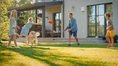 Smiling family of four tossing around frisbee in backyard together with golden retriever on a sunny day.