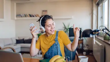 Woman wearing yellow shirt and overalls, sitting in living dancing to music while wearing headphones and holding smartphone.