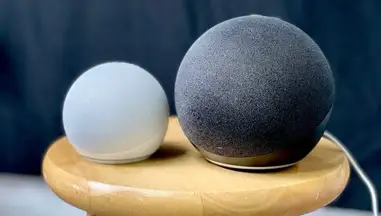 Two Echo Dot speakers, one white and one black, sitting atop a stool alongside each other.