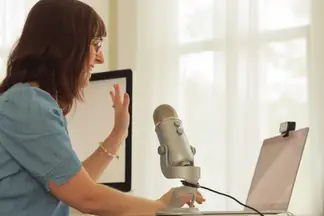 A woman waves hello at her laptop with a large USB microphone in the foreground.