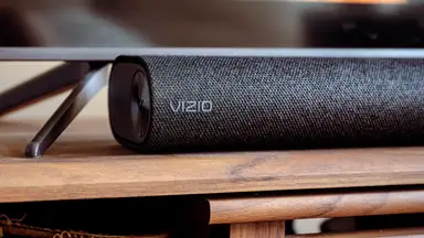 A fabric covered black Vizio soundbar sits below a TV on a wooden console.