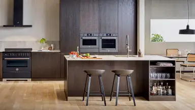 A lovely earthtone-themed kitchen with hardwood floors. There is a kitchen island, a hanging lamp, and a Bosch wall oven along the far wall.