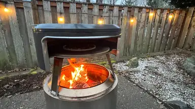 Solo Stove Pi Fire pizza oven with open flame outdoors in front of wooden fence with string lights.