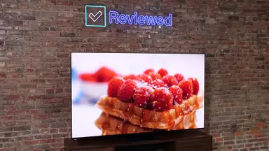 The Samsung S95C QD-OLED TV on a entertainment center and Reviewed sign about the TV, with a brick wall in the background.