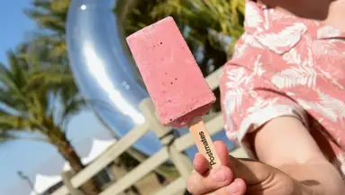 A person in a pink floral shirt holds a pink popsicle close to the camera lens.