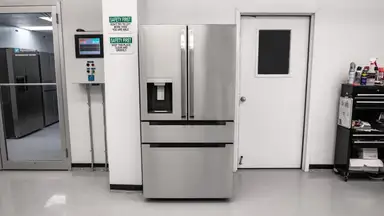 A stainless-steel refrigerator with exterior dispenser stands in a white lab environment next to a white door with a black window.