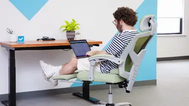Person working with a laptop on their lab while slightly reclined in the green Edx Reclining Office Chair with Footrest sitting in front of an office desk.