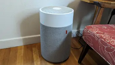Gray and white Blueair Blue Pure 311i Max purifier sitting on top of wooden floor boards next to padded chair in front of white wall.