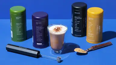 Mud/Wtr product containers next to a clear glass of a Mud/Wtr drink and a hand-held blender and spoon with Mud/Wtr product on it.