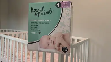 A box of Rascal and Friends size one diapers sits on the edge of a crib in a gray room