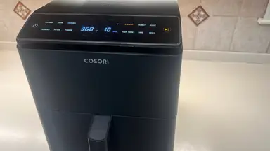 The Cosori Dual Blaze Smart air fryer on countertop in front of tiled wall.
