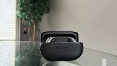 An open black charging case with hearing aids inside sits on top of a glass table with a tree in the background