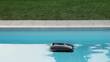 A black, square robot floats on the surface of a swimming pool
