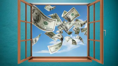 Dollar bills fly out an open window into a blue sky