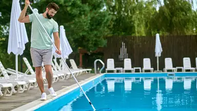 Man manually cleaning a swimming pool with a long skimmer pole beside lounge chairs and umbrellas at an outdoor pool area.