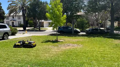 Yarbo Core Robot Lawn Mower shown mowing green grass in front of a series of trees and cars lined on the road.