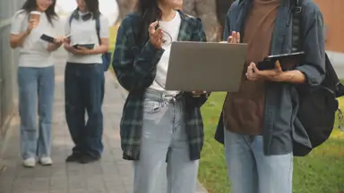 A group of students on the sidewalk holding notebooks, a laptop and an iPad