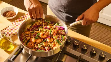 A person drizzles sauce over a skillet of shrimp, noodles, edamame, and purple cabbage stir-fry on the stovetop.