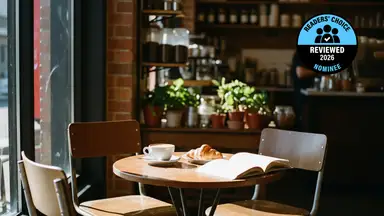 A coffee cup and croissant on a small wooden café table next to an open book.