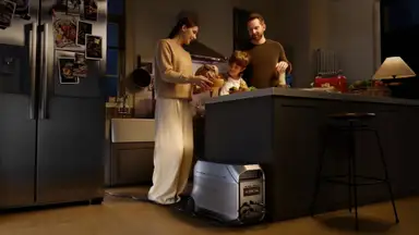 A family including a woman in a tan sweater and white pants stands next to a brunette male child and a man in a brown sweater, huddled around the kitchen counter. Their refrigerator, on the left side of the image and decorated with photos, is being powered by an EcoFlow device.