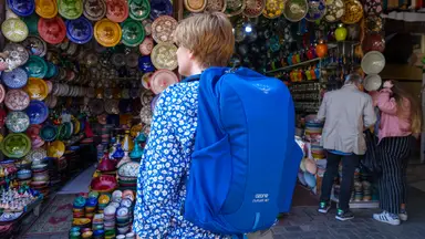 A woman wearing a blue Osprey travel backpack stands outside a pottery vendor in a Morocco market.