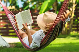 Woman wearing white dress and tan straw hat laying in multi-colored hammock outside while reading a book.