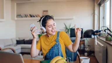 Woman wearing yellow shirt and overalls, sitting in living dancing to music while wearing headphones and holding smartphone.