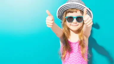 Little happy girl in beach outfit with hat and sunglasses holding thumbs up on blue background.