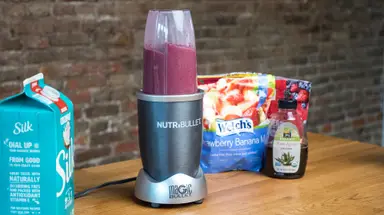 A Nutribullet personal blender filled with a berry smoothie sits on a counter next to its ingredients.